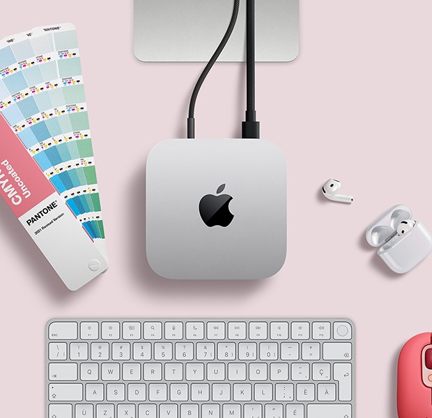Mac mini, vue en plongée, aluminium argent, coins arrondis, logo Apple noir brillant centré, branché à un câble, support du Studio Display partiellement visible, AirPods 4, nuanciers Pantone, clavier et souris Logitech rouges