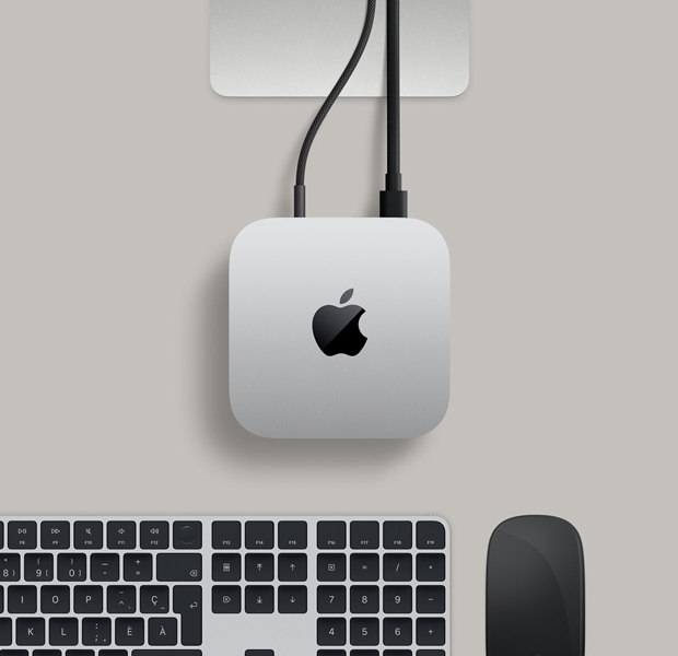 Mac mini, vue en plongée, aluminium argent, coins arrondis, logo Apple noir brillant centré, support du Studio Display partiellement visible, Magic Keyboard avec Touch ID, Magic Trackpad