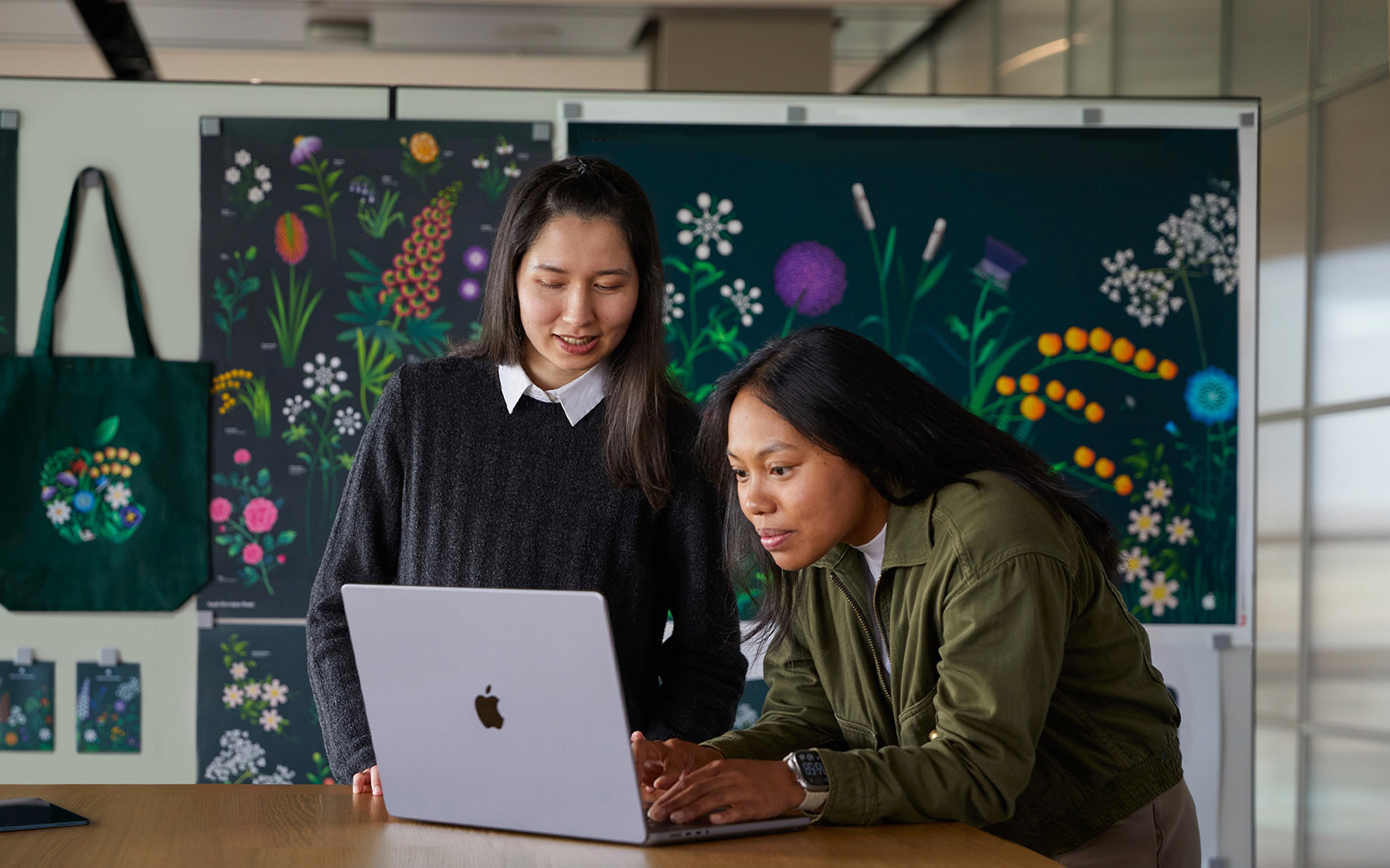 Deux membres de l’équipe Marketing d’Apple travaillent ensemble sur un MacBook devant des affiches conçues par leur équipe.
