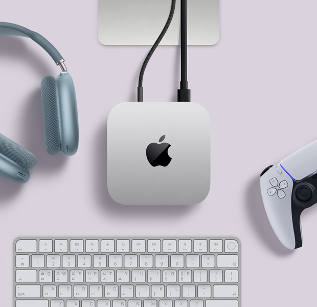 Mac mini를 위에서 바라본 모습. 실버 알루미늄 외장, 둥근 형태의 모서리, 중앙의 광택 나는 검은색 Apple 로고가 보이며, 코드가 연결되어 있습니다. Studio Display의 스탠드 일부가 보이며, 블루 색상 AirPods Max 2, 화이트 색상 PlayStation DualSense 컨트롤러, 화이트 색상 키보드가 놓여 있습니다.
