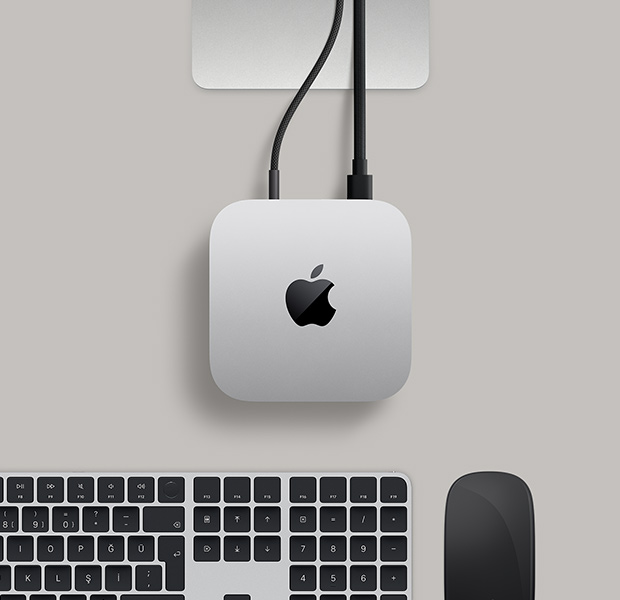 Mac mini, üstten görünüm, gümüş rengi alüminyum, yuvarlatılmış köşeler, ortalanmış parlak siyah Apple logosu, Studio Display standının kısmi görünümü, Touch ID özellikli Magic Keyboard, Magic Trackpad