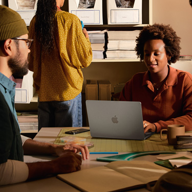 Colegas a colaborar juntos numa secretária. Na secretária estão um tapete de corte e outras ferramentas de design. Um colega interage com um MacBook.