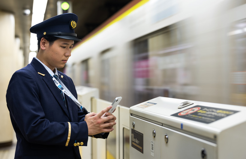 En stationsmedarbejder står på perronen og bruger sin iPhone til at scanne en QR-kode, mens et tog kører forbi.
