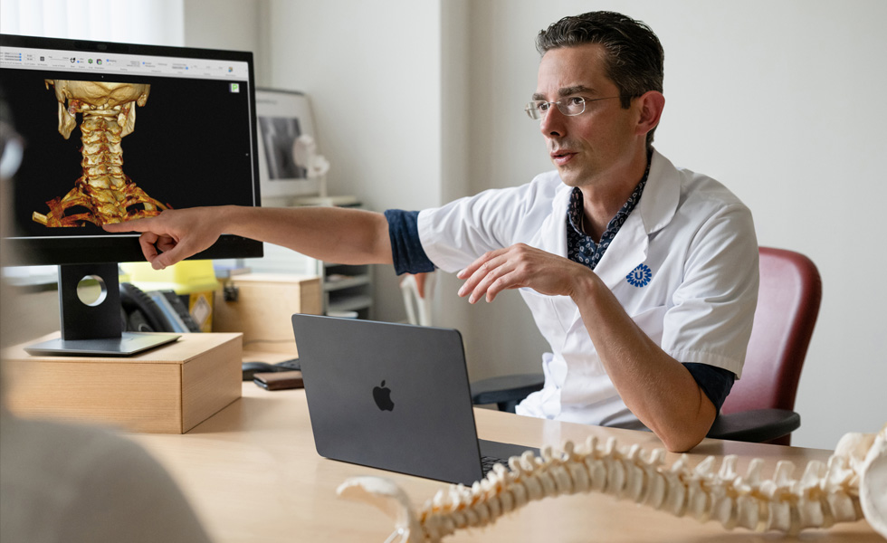 En læge peger på en 3D-gengivelse af en nakkes skelet på en Studio Display, mens en MacBook står på skrivebordet