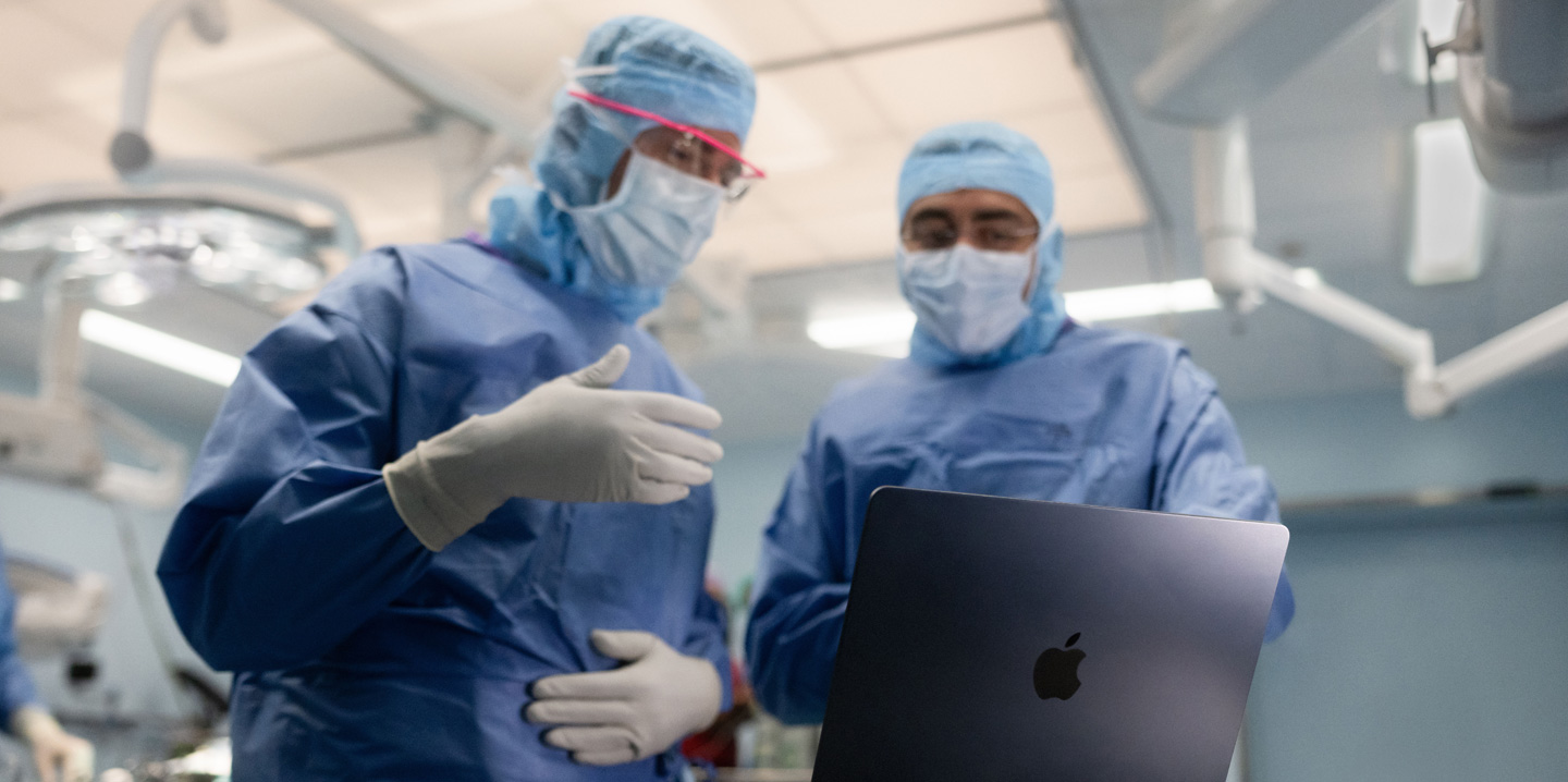 To kirurger på operationsstuen ser på en MacBook, og den ene gestikulerer med hånden, mens den anden ser på